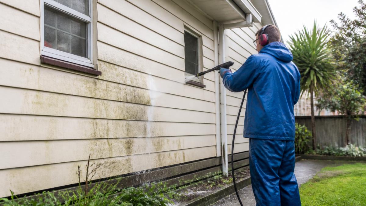 A professional house washer in safety gear using a high-pressure hose on the exterior wall of a suburban home, with clean water runoff visible and a garden nearby