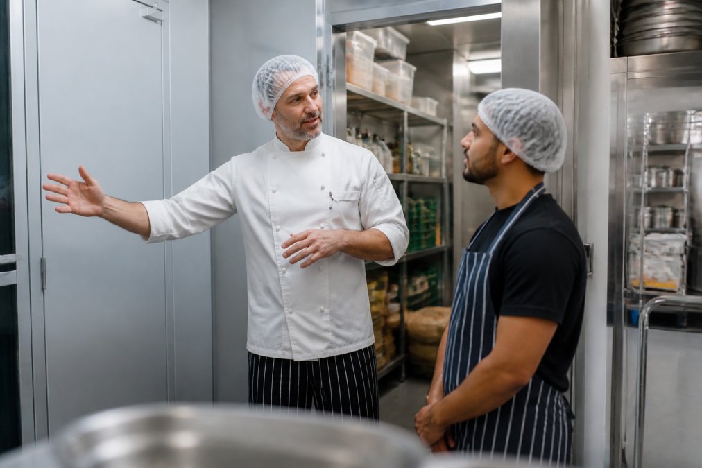 A head chef in a white uniform showing a new temporary kitchen worker where the walk-in fridge and dry store are, both wearing hairnets, stainless steel kitchen in the background