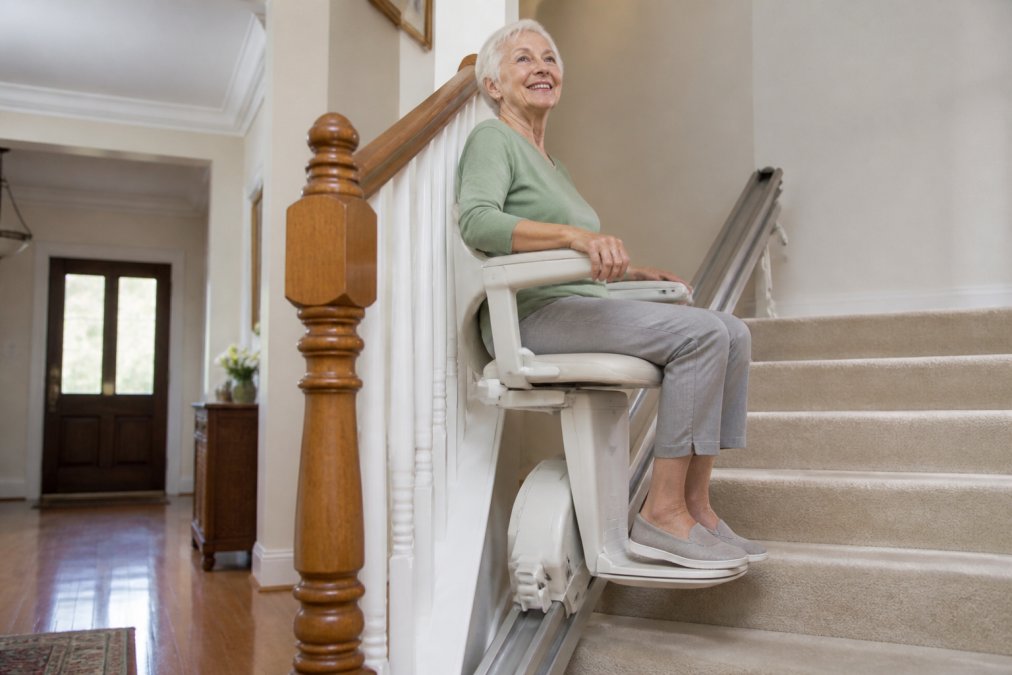 Older adult confidently ascending home staircase using stairlift with visible seat, safety features, and secure grip on armrest in bright residential setting