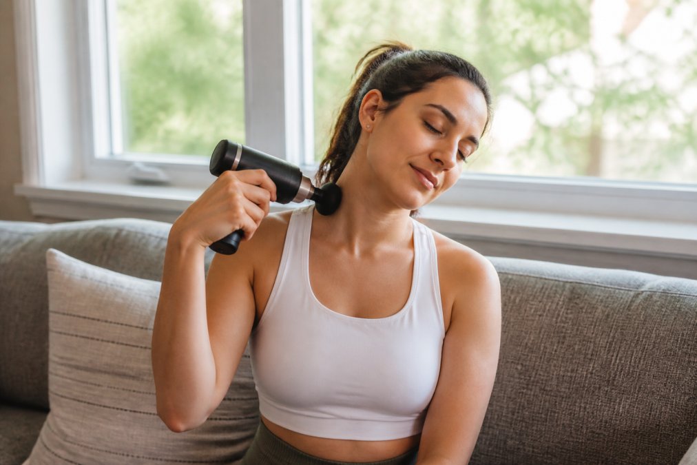 Person sitting on a couch using a handheld percussion massage gun on their shoulder and neck area, relaxed posture with natural window lighting in background