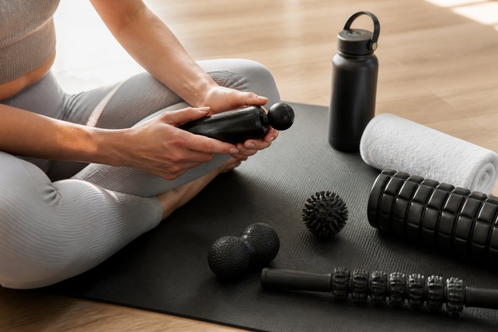 Close-up of hands holding a massage tool while sitting on a yoga mat with water bottle and rolled towel nearby in natural lighting