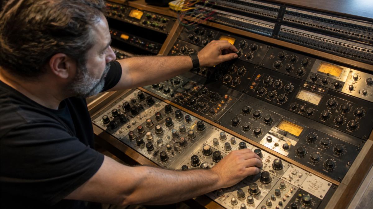 Professional audio engineer adjusting analog compressor and EQ on mastering console with patchbay and audio interface connections visible in modern mastering studio