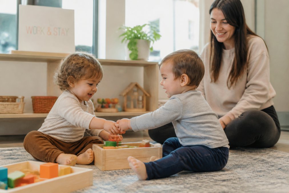 Two toddlers playing together with toys, smiling and interacting with genuine engagement while a caregiver supervises nearby in a colorful, safe play environment