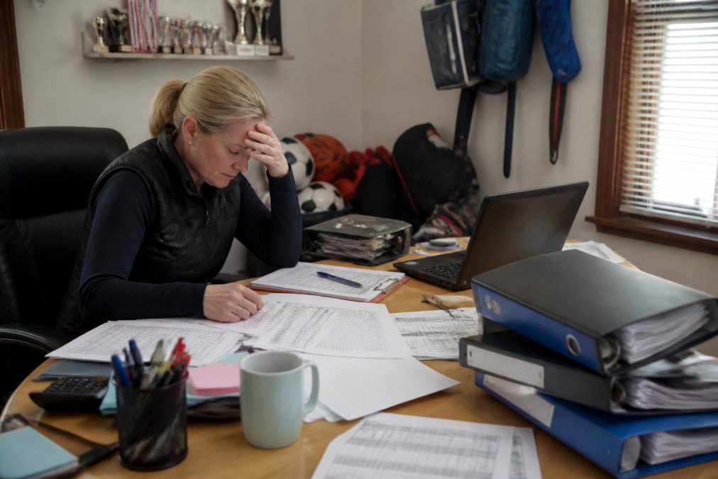 A stressed volunteer club administrator looking at multiple monitors and paper spreadsheets in a cluttered office, with sports equipment in the background