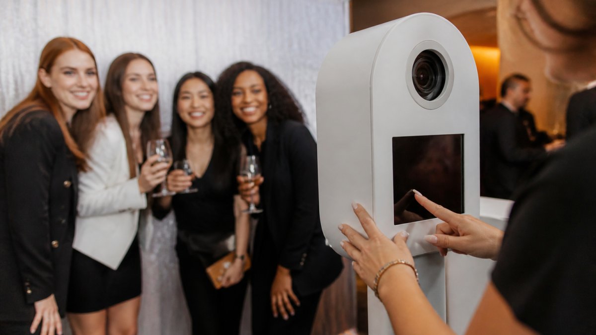 Guests smiling and posing in front of a branded photo booth at a corporate event, with custom backdrop featuring company logo and colors, professional attendant managing the experience in modern venue with warm lighting