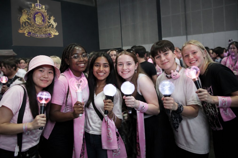 Diverse group of K-Pop fans wearing official fan club merchandise and holding light sticks in group colours, gathered together at a fan event, smiling and enjoying the atmosphere