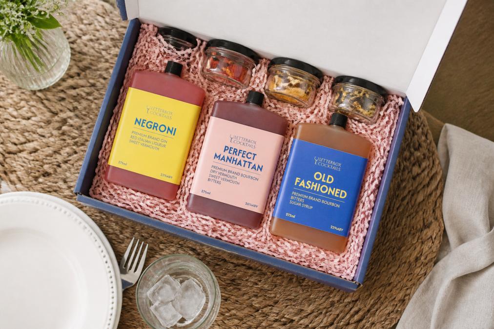 Overhead view of an open bespoke cocktail gift box displaying multiple premium bottles with hand-crafted garnishes and elegant presentation on a dining table, soft natural lighting from a window
