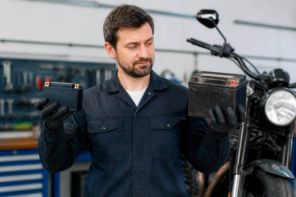 A motorcycle mechanic holding a lithium motorcycle battery in one hand and a lead-acid battery in the other, showing the clear weight and size difference in a bright workshop