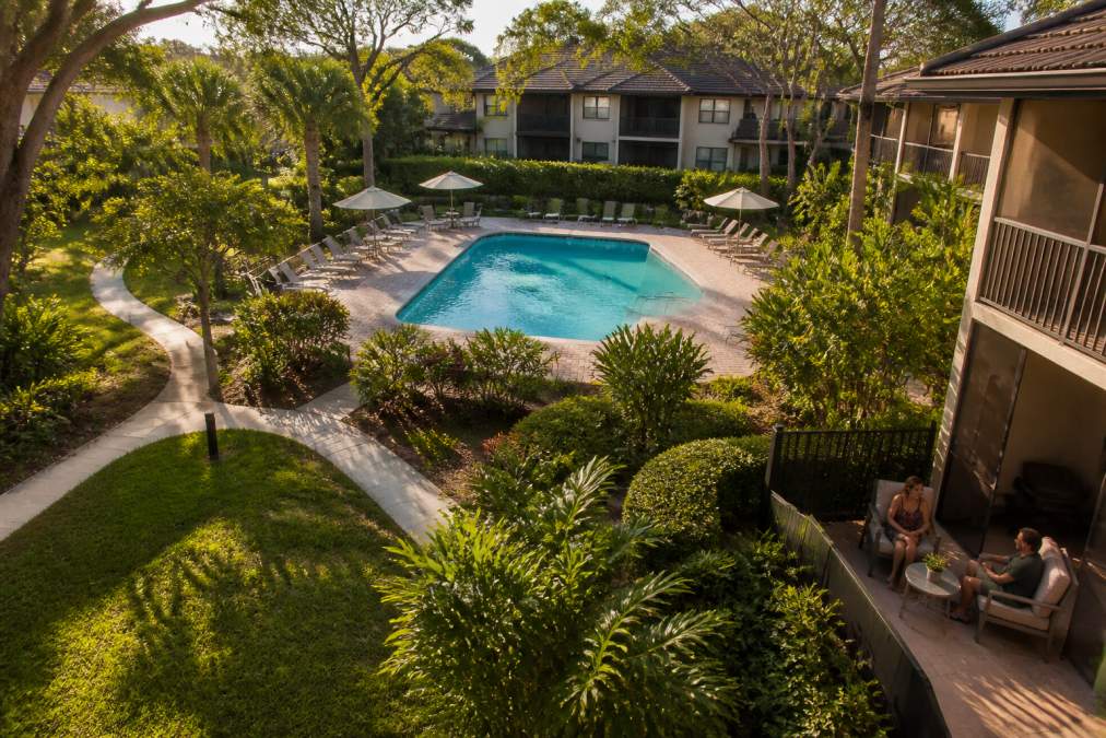 Benefits of Living in Garden Style Apartments 1 Aerial view of a serene garden-style apartment community with low-rise buildings, lush green landscaping, residents relaxing on private patios near a resort-style pool, afternoon sunlight filtering through mature trees