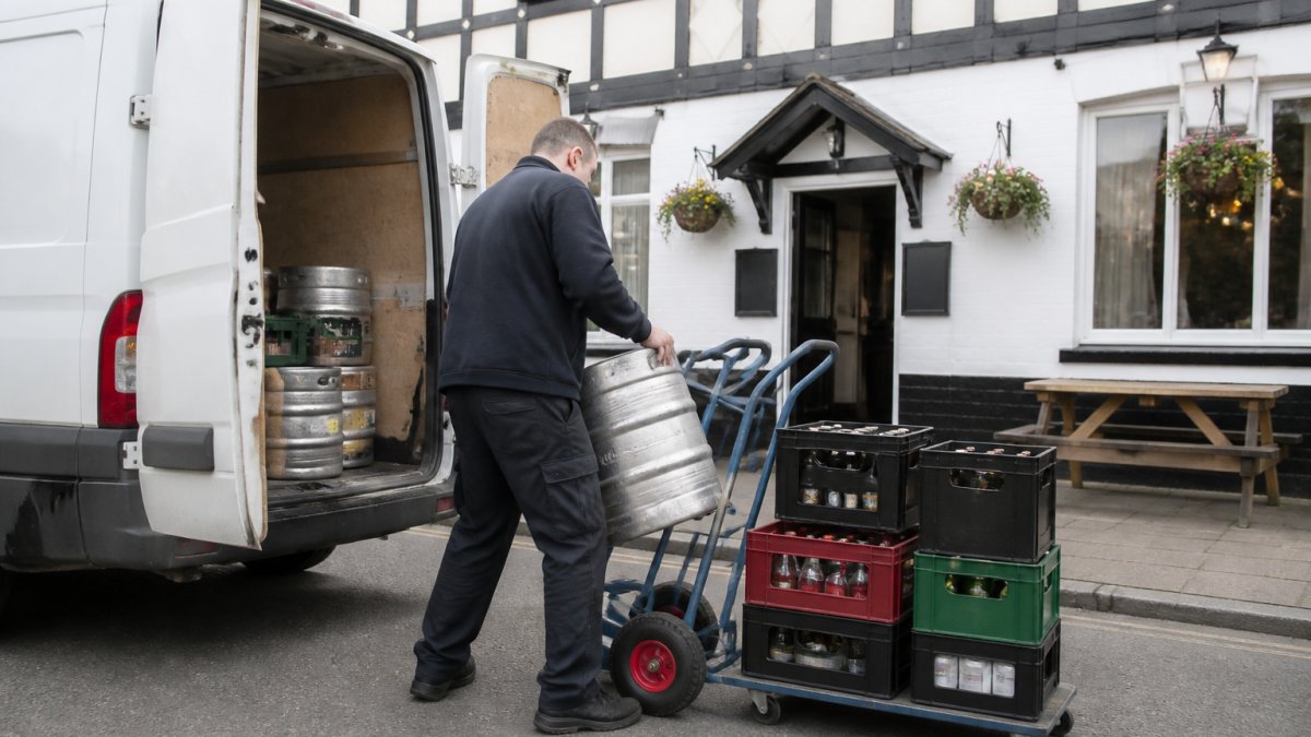 A white delivery van parked outside a traditional British pub, with a worker in a plain uniform unloading kegs and crates of drinks onto a trolley in daylight