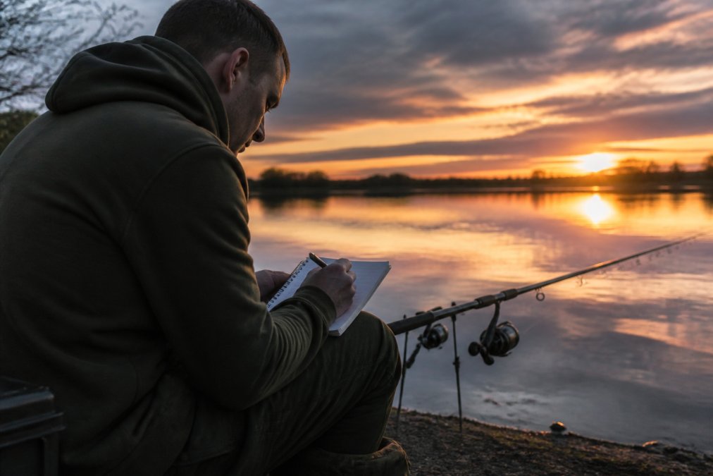 Angler sat by the waterside at dusk, holding a smartphone and notepad, recording session details with the water and rod visible in the background