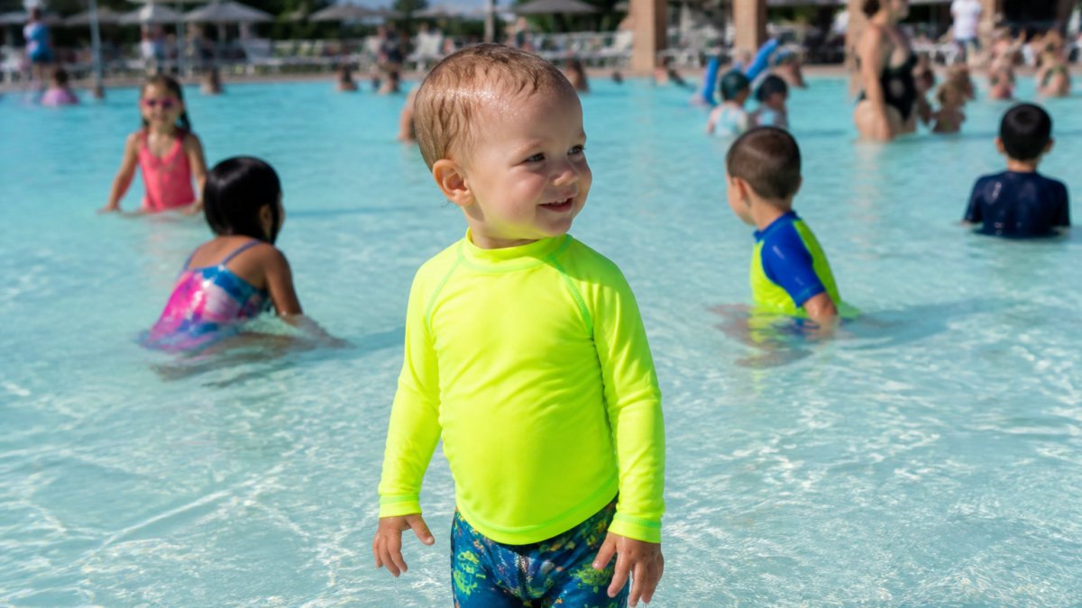Toddler in bright neon long sleeve swim shirt standing in shallow water at a busy public pool, surrounded by other children, the vibrant color clearly visible against the blue water and pool deck