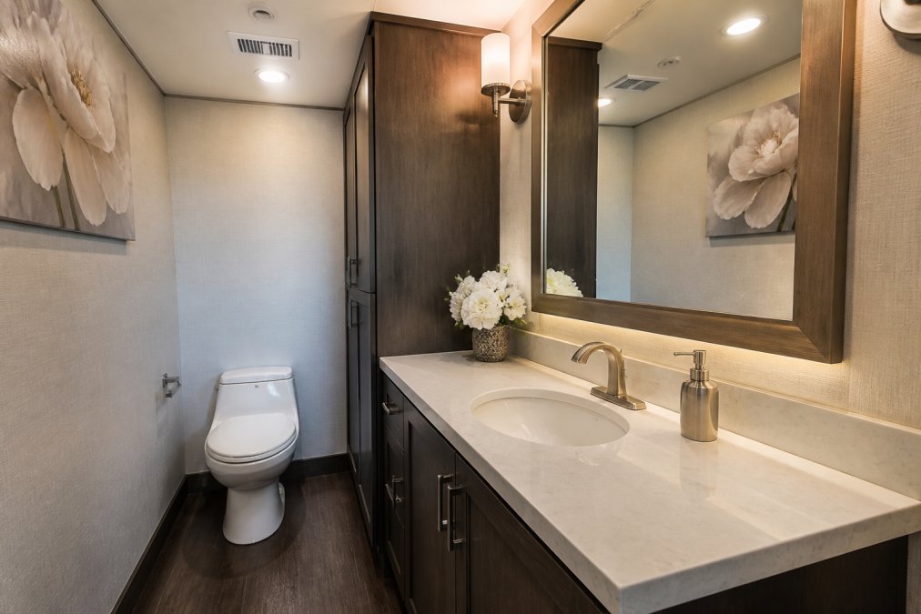 Interior of a luxury restroom trailer with a flushable toilet, sink with running water, vanity mirror, and warm interior lighting