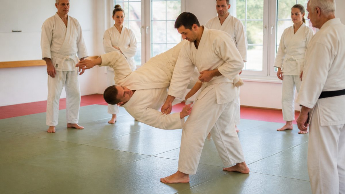 A small group of adult students in white uniforms practicing a controlled throwing technique on a padded mat in a bright dojo, instructor watching closely