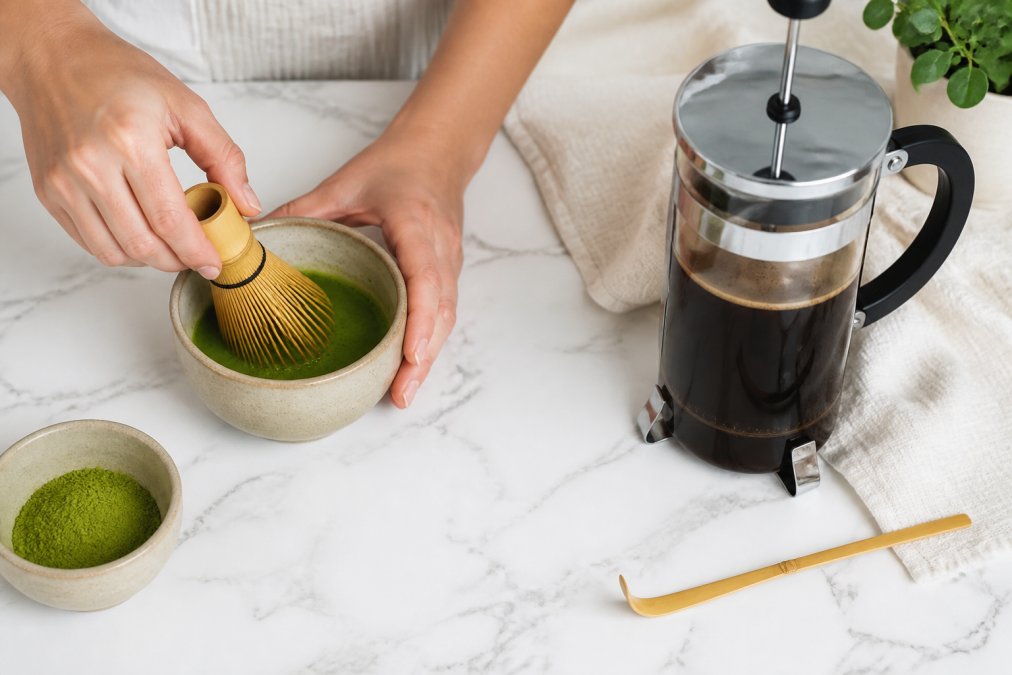 Overhead shot of hands whisking bright green matcha powder in a small bowl next to a French press filled with dark coffee, on a clean white kitchen counter with a bamboo whisk
