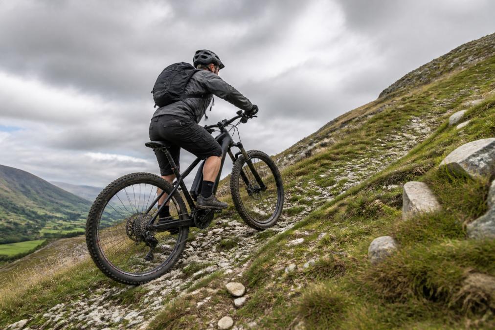 A cyclist riding a mid-drive electric bike up a steep countryside hill, motor visible at the cranks, rider pedalling in a low gear under overcast light