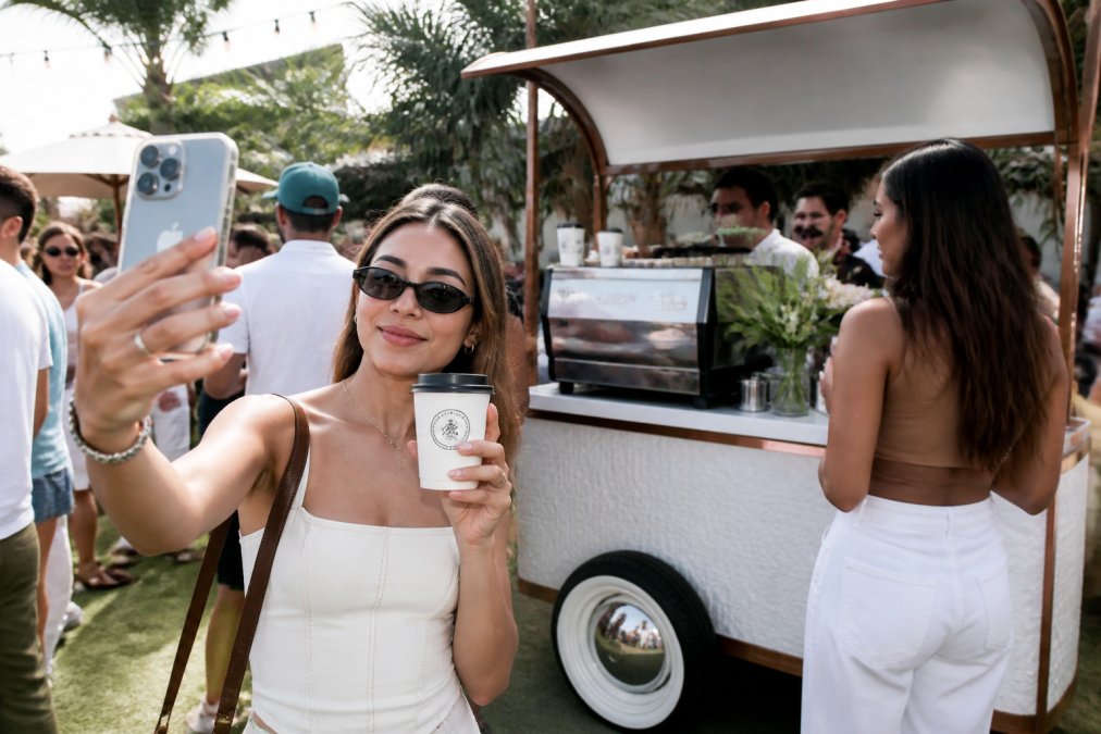 Guest holding a custom-branded coffee cup with Colada Kulture Co. logo, taking a selfie with the decorated mobile coffee cart and other attendees visible in the background, natural outdoor event lighting