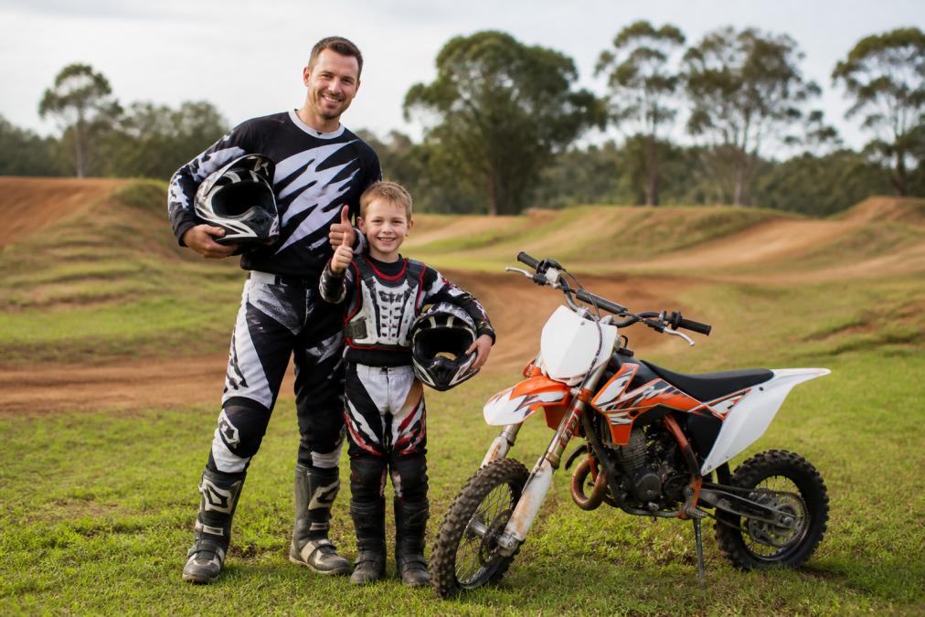 A parent and young child in full motocross gear standing next to a small dirt bike on a grassy track, smiling and giving a thumbs up, with helmets under their arms