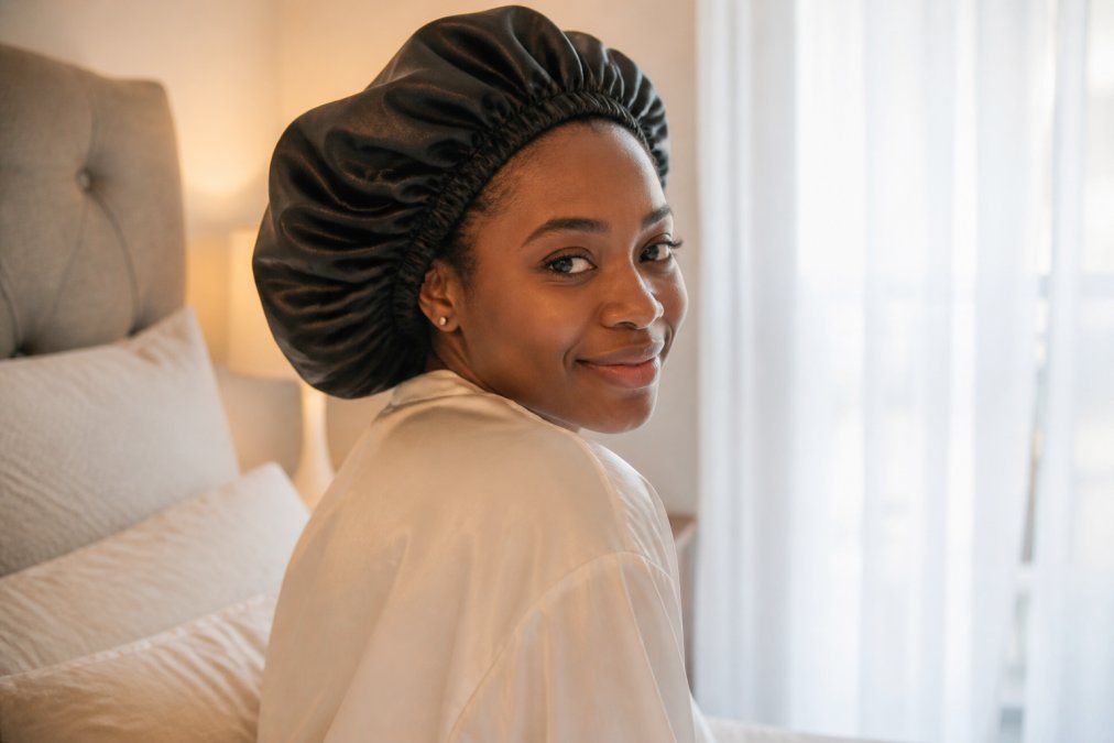 Close-up photograph of a Black woman with coily natural hair wearing a luxury mulberry silk bonnet in a softly lit bedroom, morning light through sheer curtains, serene and elegant mood
