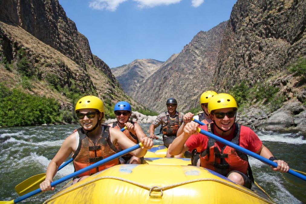 A group of smiling rafters paddling through a scenic canyon river with towering rock walls on both sides, wearing helmets and personal flotation devices, under bright afternoon sunlight