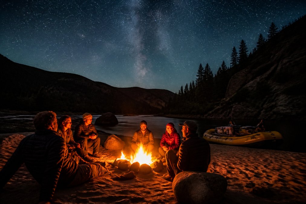 A group of friends gathered around a campfire on a sandy riverbank at dusk, with a raft visible in the background and a star-filled sky overhead, warm firelight illuminating their faces