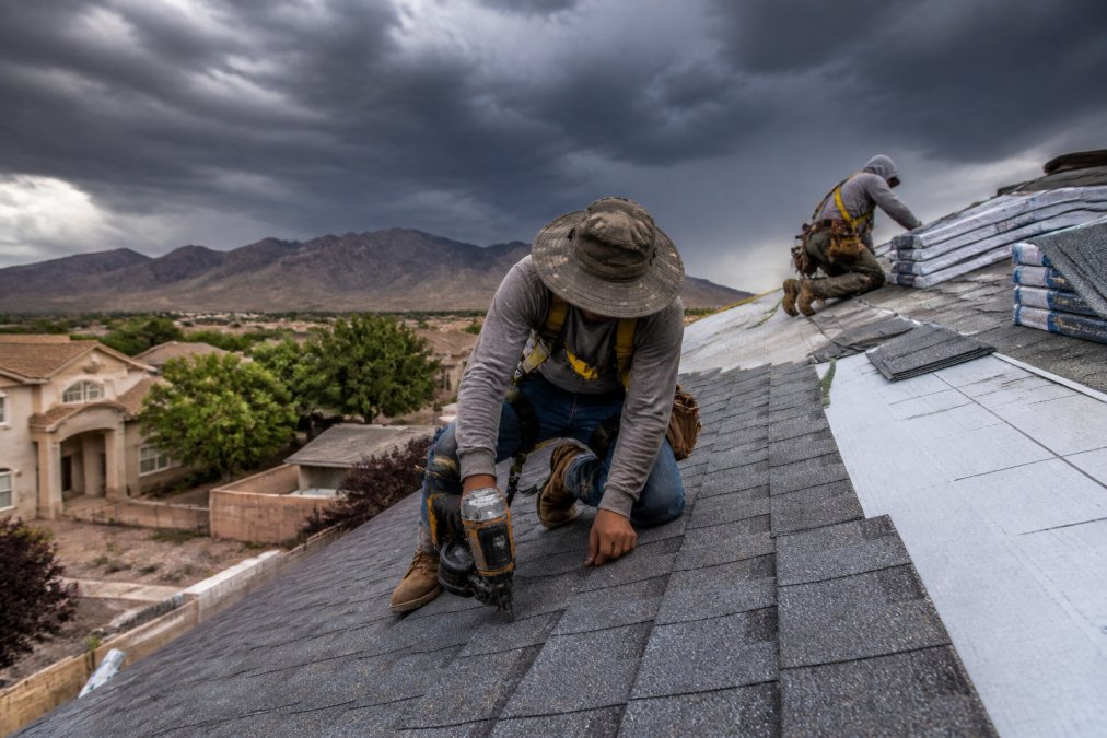 Roofing crew installing new shingles on a residential roof in El Paso with dark monsoon clouds building in the background, workers in safety gear securing materials