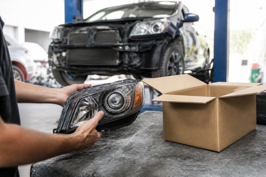 Close-up of a mechanic's hands holding a genuine OEM car part next to its packaging, with a vehicle on a lift in the background of a clean repair shop