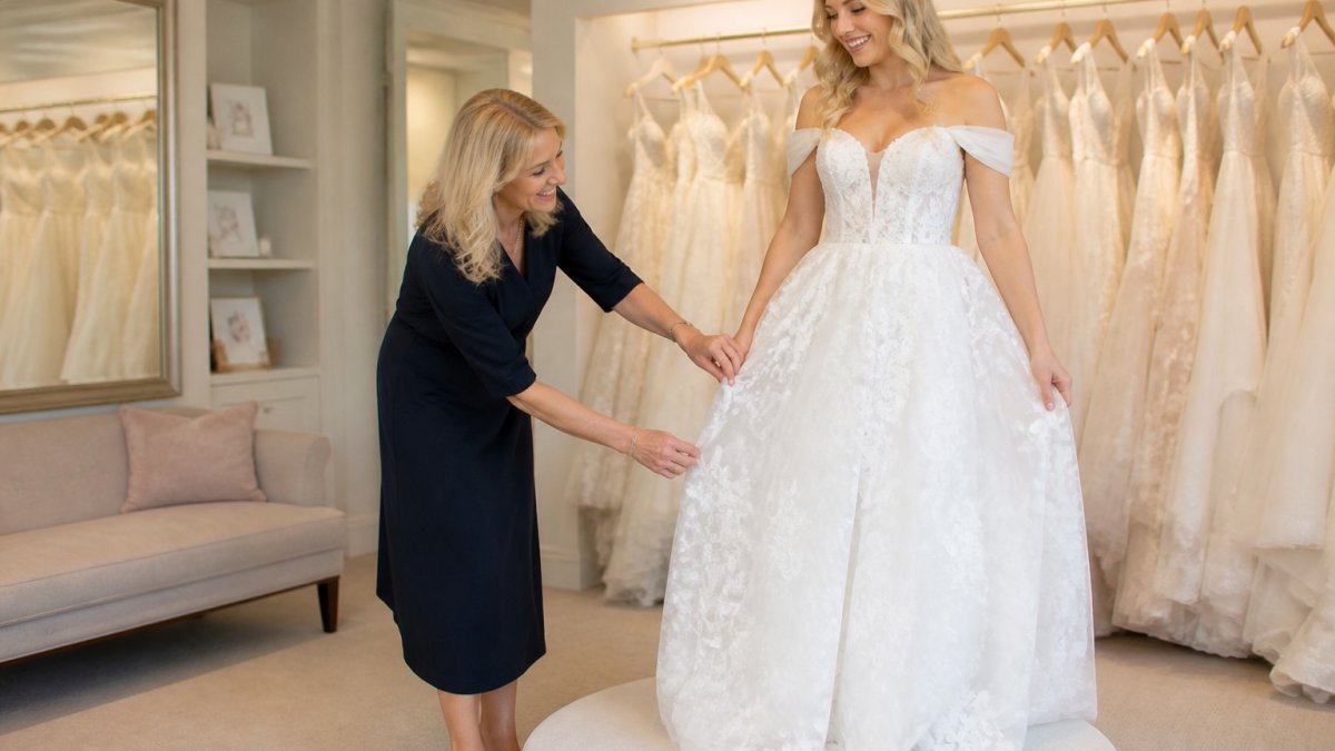 A bridal stylist in a bright boutique helping a smiling bride step into a wedding dress on a fitting pedestal, with a rail of gowns softly blurred in the background