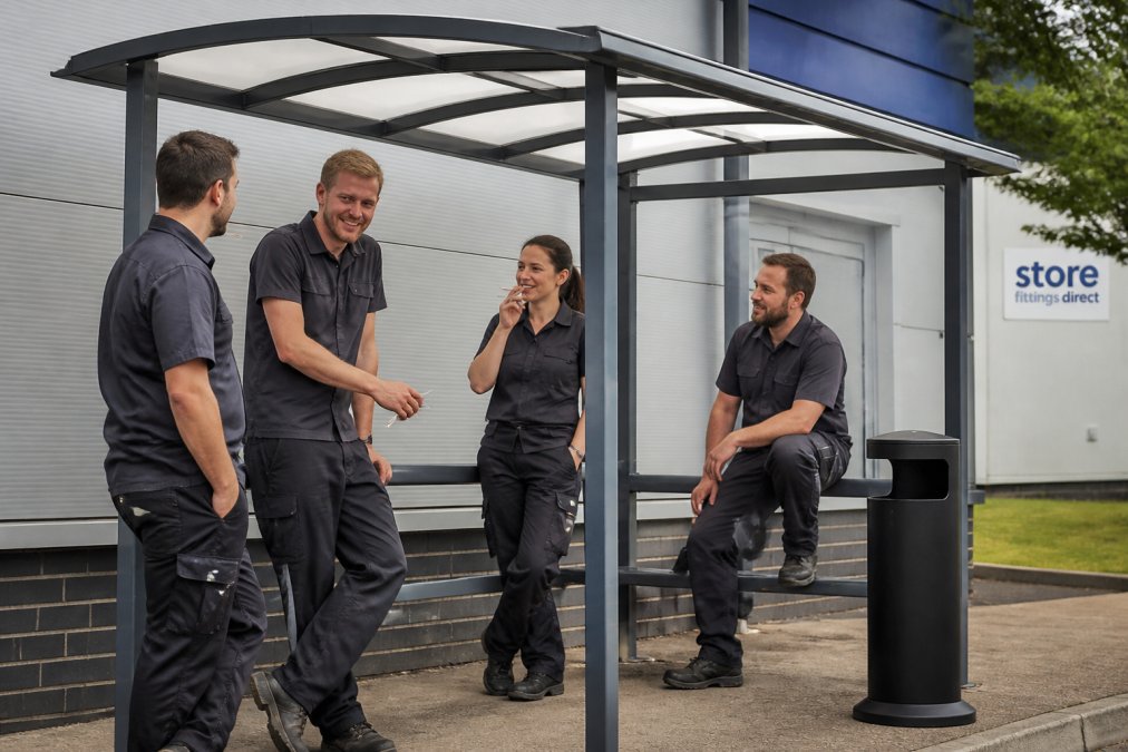 Employees chatting and relaxing under a modern outdoor smoking shelter on a bright day outside a commercial building.