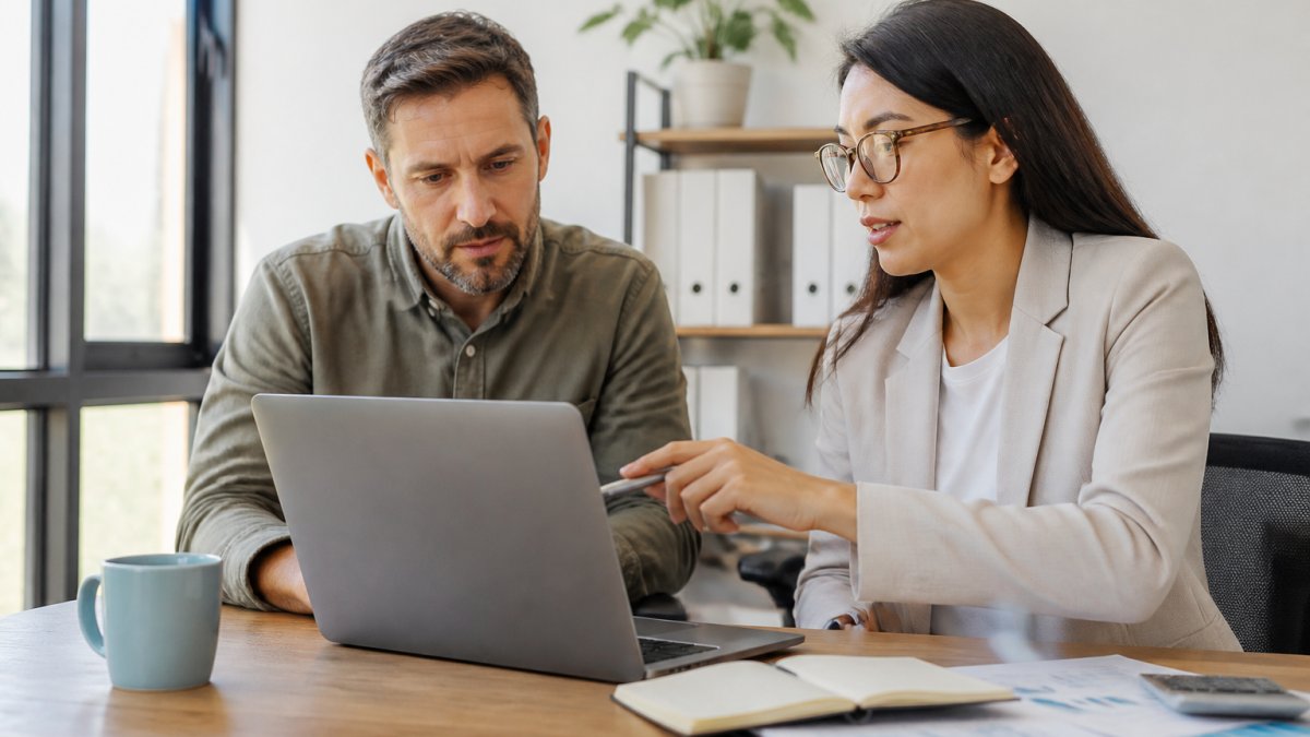 A small business owner reviewing financial statements on a laptop in a bright office while a professional accountant points at the screen and explains the numbers