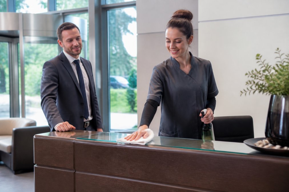 A professional cleaner in a branded uniform polishing a reception desk in a bright, modern office lobby while a manager observes approvingly