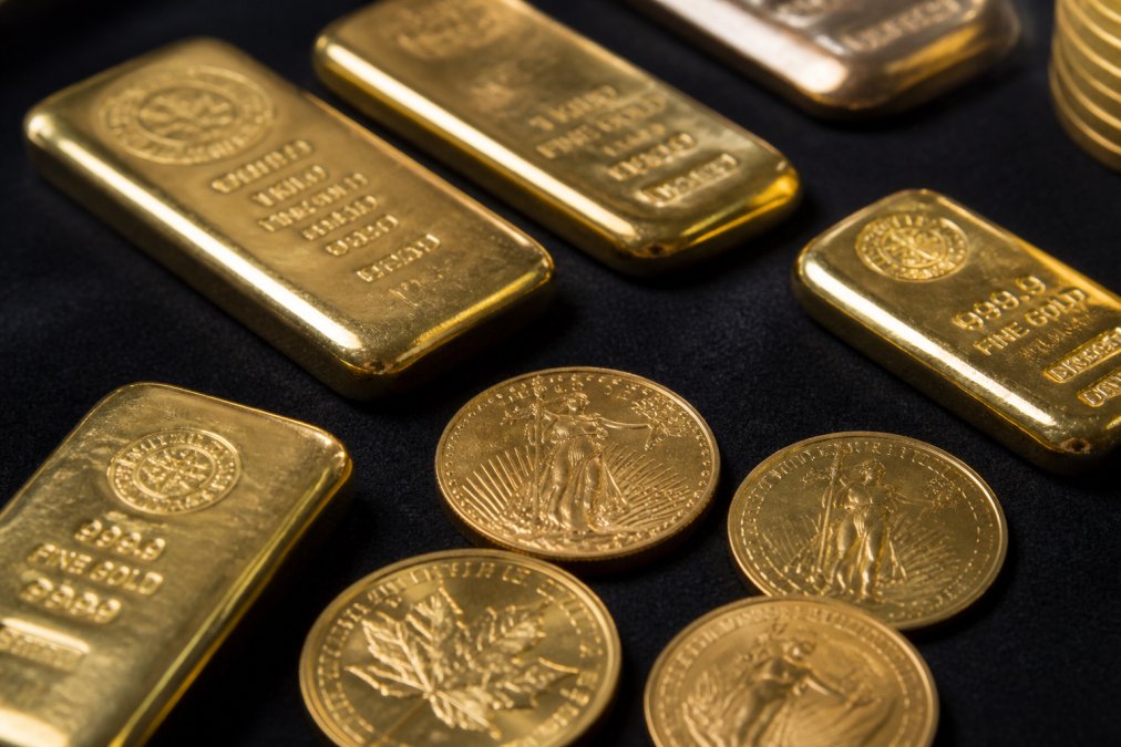 Close-up of several gold bullion bars and coins arranged on a dark felt surface, soft natural light catching the mint marks and stamped purity
