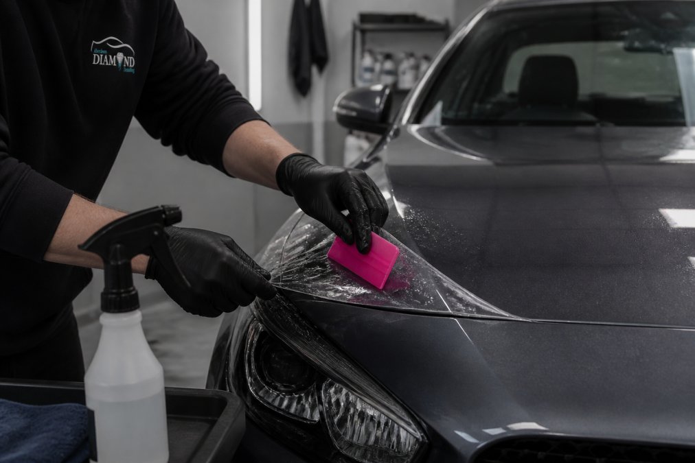 Close-up of a professional detailer's gloved hands applying transparent paint protection film to a car bonnet edge, squeegee and spray bottle visible, clean detailing studio lighting