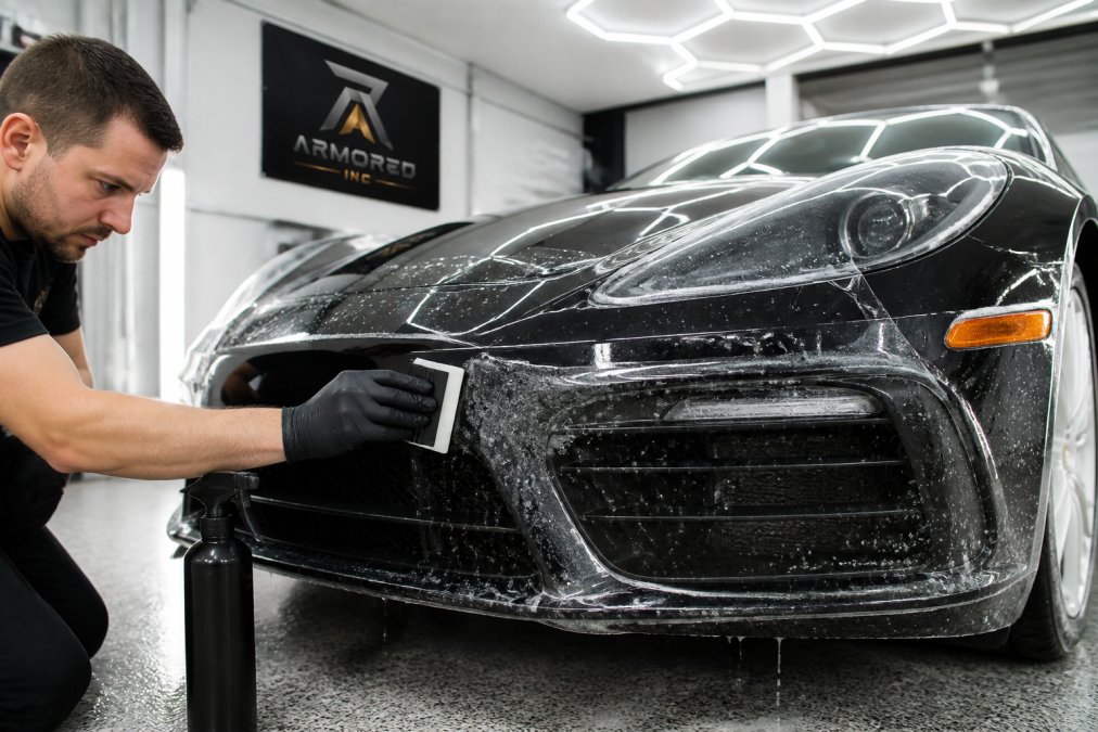 Professional detailer applying a clear film to the front bumper of a luxury sports car inside a bright, clean detailing studio, using a squeegee to smooth the film