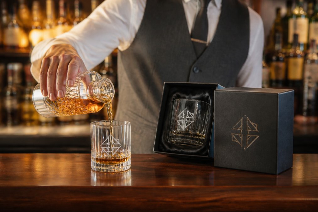 Professional pouring whiskey into an etched tumbler on a polished wooden bar counter, soft amber lighting highlighting the engraved logo detail
