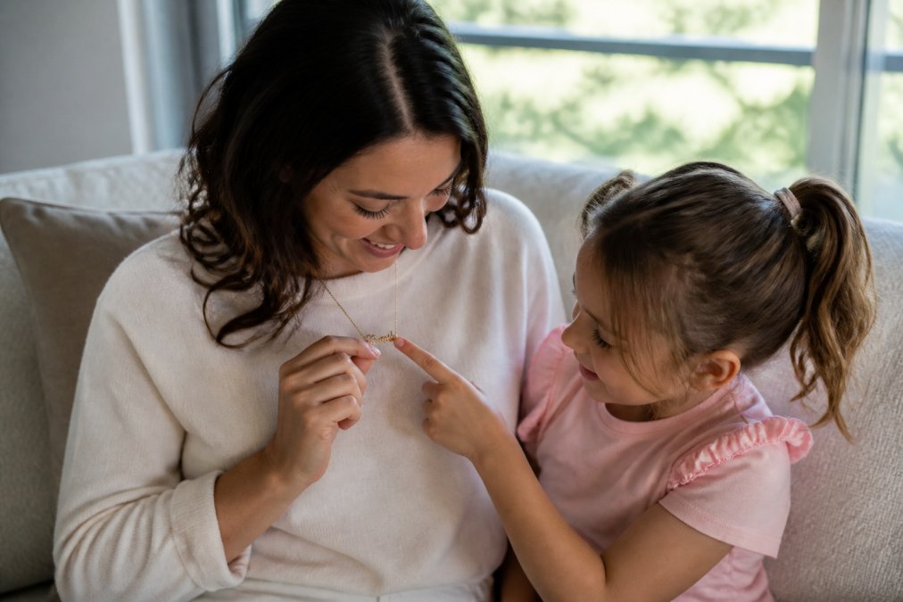 A godparent and young child sitting together on a sunlit couch, the godparent gently holding a personalized name necklace as the child points to the engraved letters