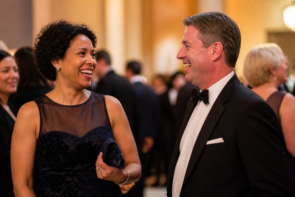A candid shot of a nonprofit executive director laughing with a donor at an elegant fundraising gala, both in formal attire, warm ambient lighting, other guests softly blurred in the background
