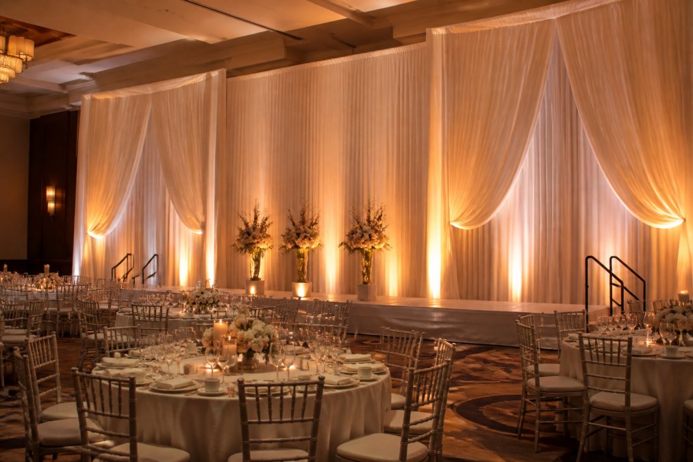 A large ballroom transformed with floor-to-ceiling white and ivory drape panels framing a central stage, warm amber uplighting casting soft shadows across pleated fabric, round banquet tables set with glassware below