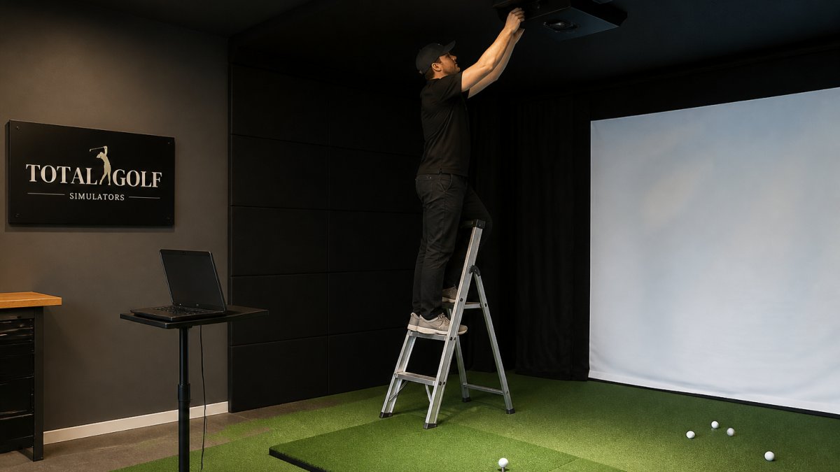 A technician on a small ladder adjusting an overhead launch monitor above a golf hitting mat, laptop open on a stand nearby showing setup software
