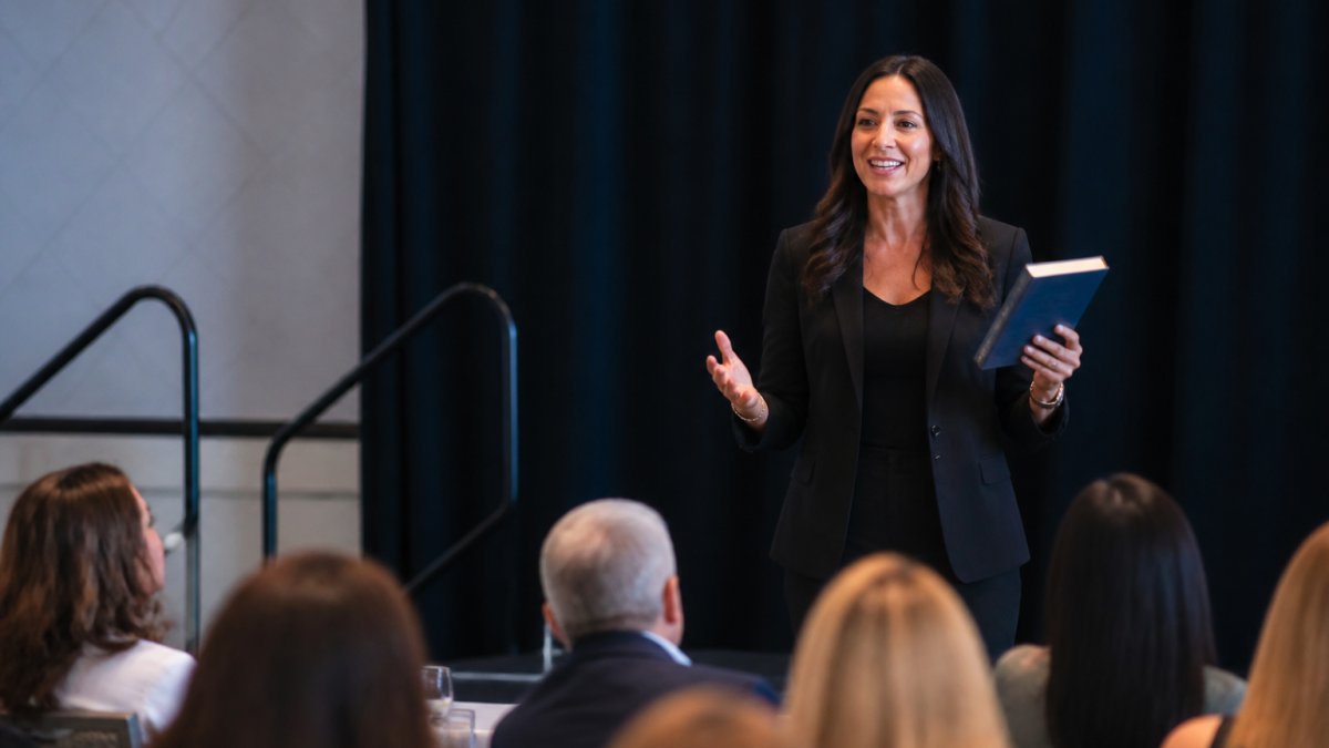 A professional coach speaking on stage at a business conference, holding a copy of their book while addressing an engaged audience