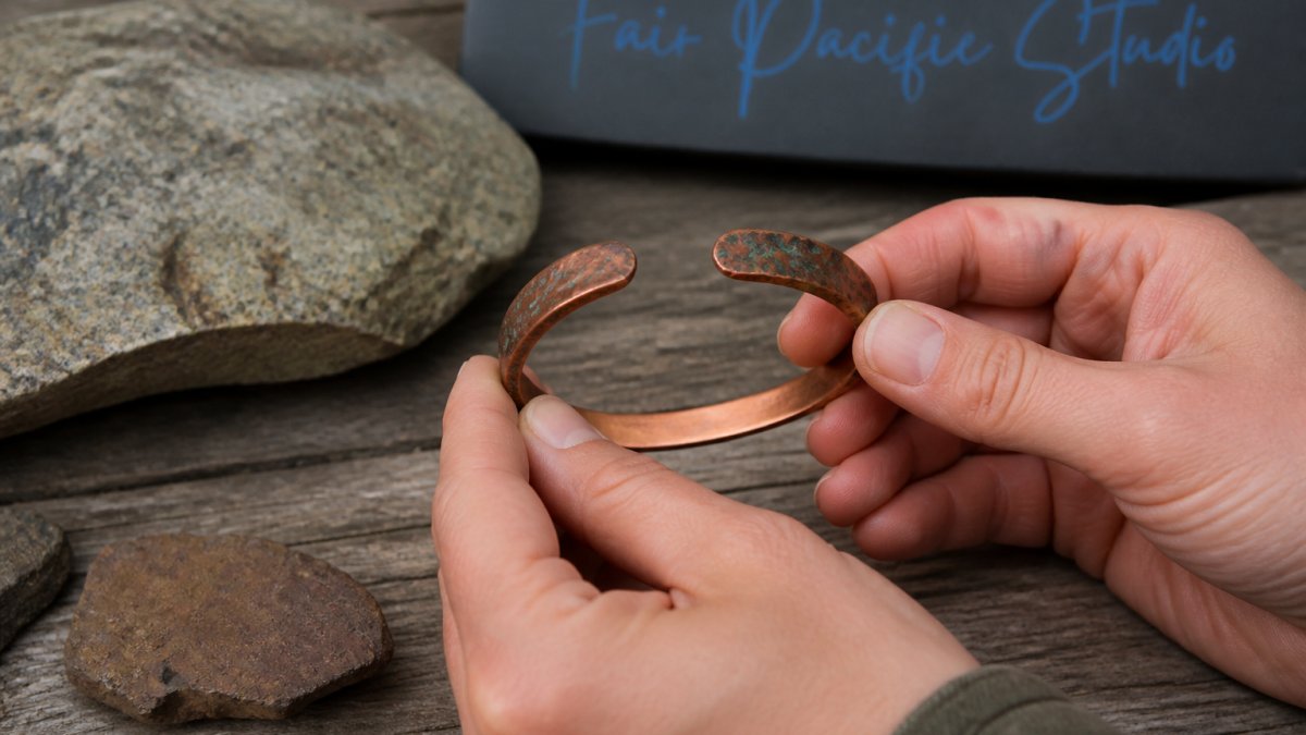 Hands holding a handcrafted recycled copper jewelry bracelet with a natural patina against weathered wood and stone.