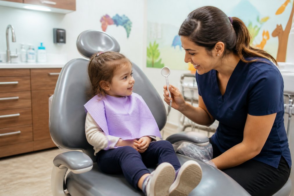 A toddler sitting comfortably in a dental chair while a friendly dental hygienist shows a small mirror, in a bright, child-friendly clinic room with colorful wall decorations