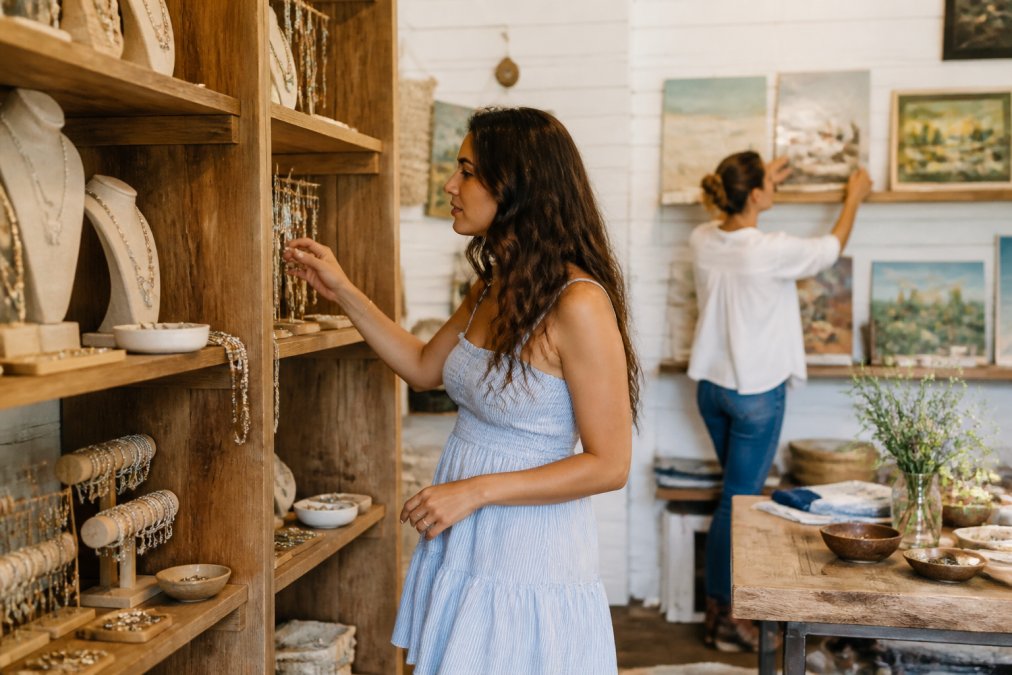 Customer browsing handcrafted jewelry displayed on elegant wooden shelving in a warm-toned boutique interior, with attentive shop staff member nearby arranging art pieces on the wall