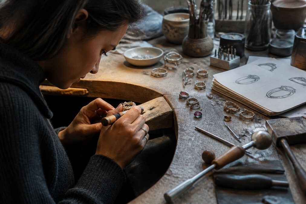 Artisan jeweler's hands carefully working on a custom piece at a wooden workbench, surrounded by gemstones, metal tools, and sketches under warm workshop lighting