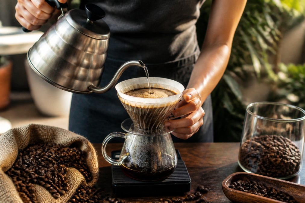 Hands pouring hot water over single origin coffee grounds in a pour-over dripper, with fresh beans and a timer visible on a wooden surface in natural light