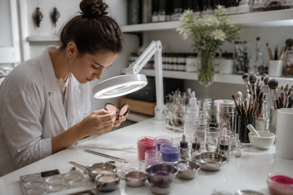 A formulator in a bright studio carefully inspecting a cosmetic compact under a magnifying lamp, with pigment powders and small mixing tools on the workbench