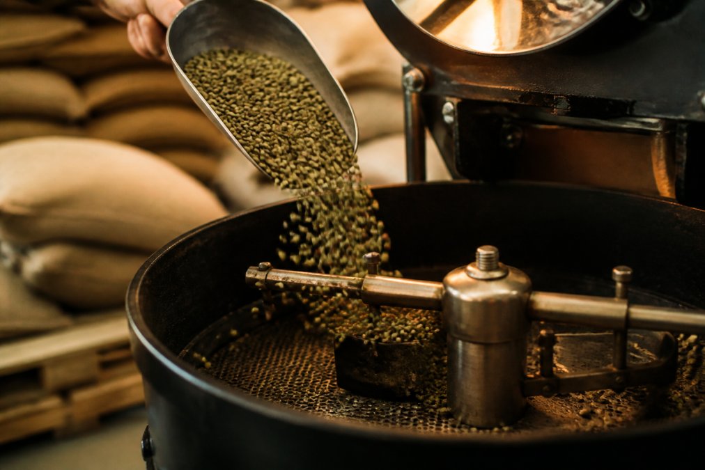 Close-up of a small coffee roaster drum with green beans cascading in from a scoop, warm amber lighting illuminating the craft roastery workspace with burlap sacks of green beans stacked in the background