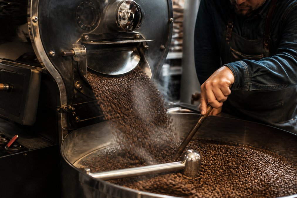 Close-up of freshly roasted coffee beans tumbling out of a small drum roaster into a cooling tray, with steam rising and a roaster's hand stirring the beans