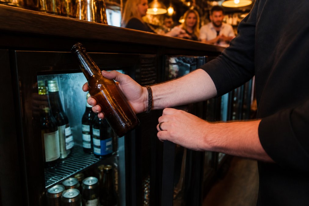 A bartender's hands lifting a chilled alcohol-free lager bottle from an under-counter fridge in a busy pub, warm amber light from the bar behind, condensation beading on the glass