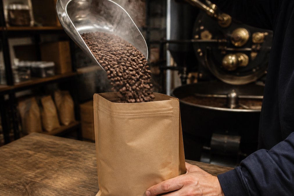 Close-up of freshly roasted coffee beans being poured into a kraft paper bag with roast date label visible, steam rising from the beans, with a coffee roastery and roasting equipment in soft background lighting